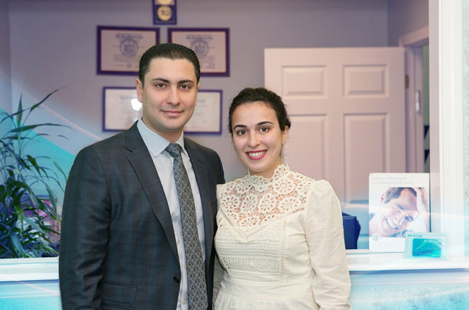The image shows a man and a woman posing together in an indoor setting, likely a professional office environment, with the man dressed in formal attire and the woman in a light-colored dress.