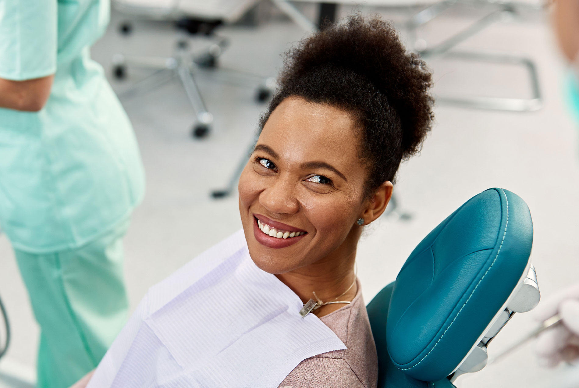 A smiling woman in a dental office, seated in a dental chair with a hygienist s station and equipment behind her.