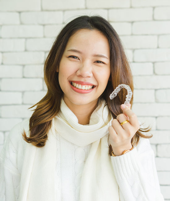 The image shows a smiling woman holding a dental impression tray, with the tray partially filled and held up to her mouth. She is wearing a white scarf and has dark hair. The background is blurred but suggests an indoor setting with a brick wall.