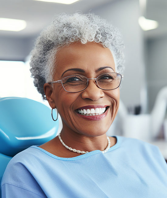 A smiling elderly woman wearing glasses, sitting in a dental chair.