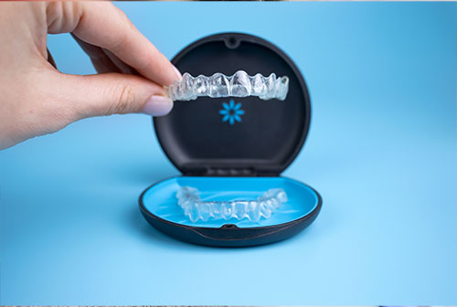 An image featuring a hand holding a clear tray containing transparent aligners, with one of the aligners placed on a black, circular object that appears to be a mouthguard or retainer case. The background is blue and minimalistic.